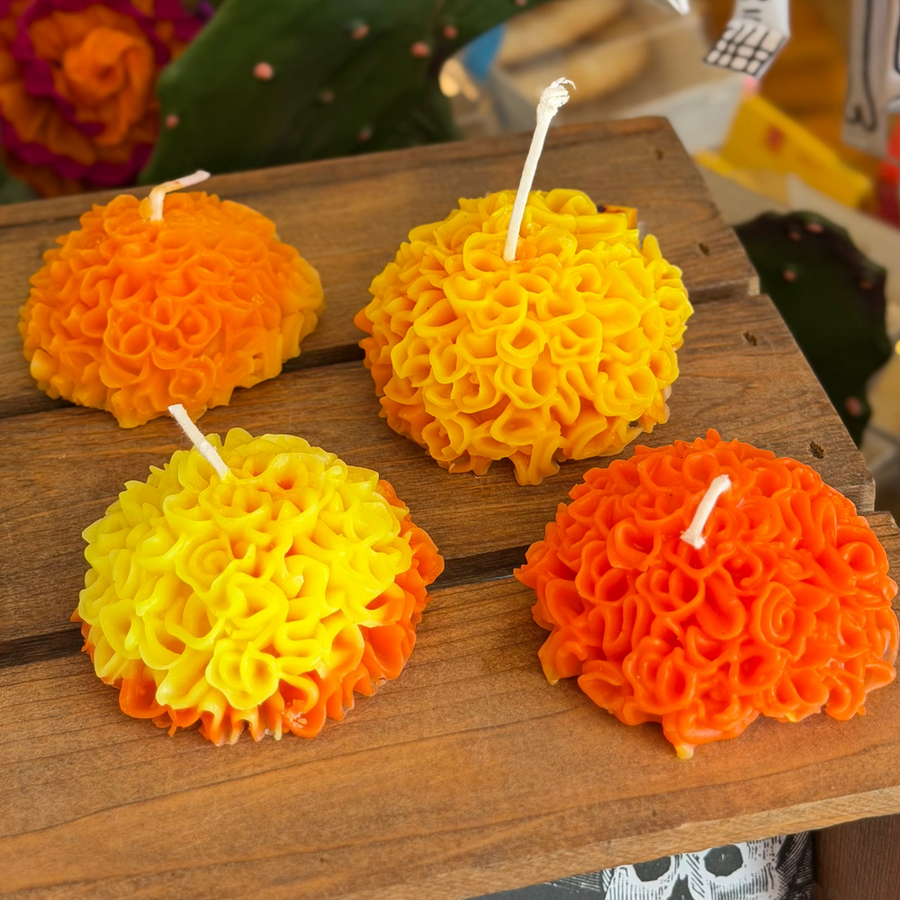 Four textured marigold candles in orange, yellow, and gradient orange-yellow combinations, arranged on a rustic wooden table with a Day of the Dead themed background.