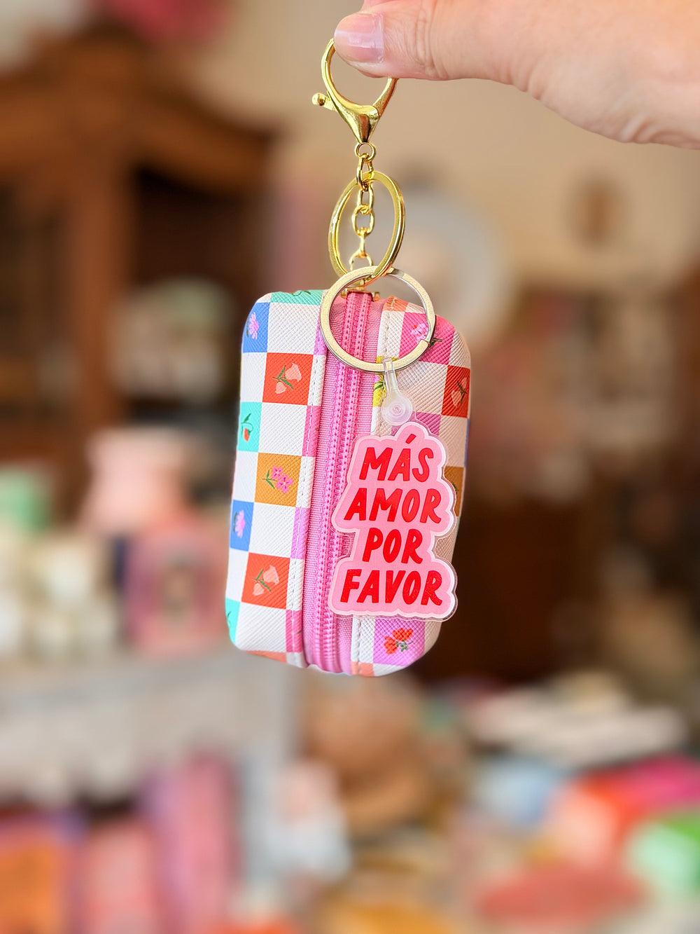 A hand holds a small zippered pouch keychain with a colorful checkered pattern, gold-tone clasp, and a pink tag that reads 'MÁS AMOR POR FAVOR' against a blurred shop background.