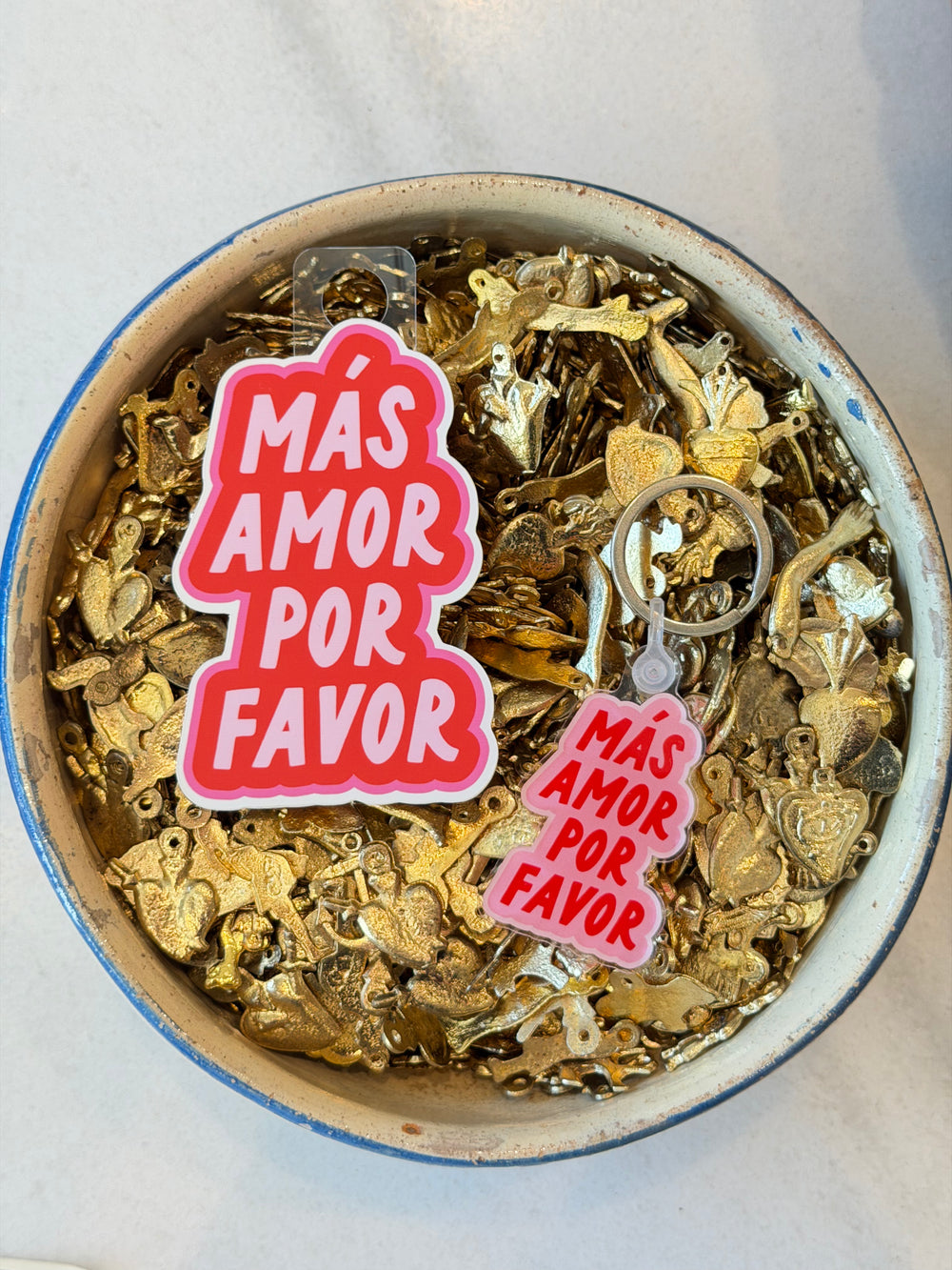 Two pink and red acrylic keychains with the Spanish text '¡MÁS AMOR POR FAVOR' resting in a bowl filled with gold-colored milagro charms on a white surface.