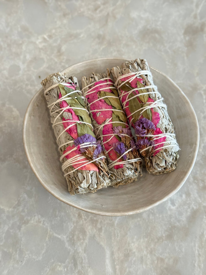 Three sage smudge sticks wrapped with pink rose petals, purple statice flowers, and green leaves, tied with white string in a bowl sitting on a marble surface.