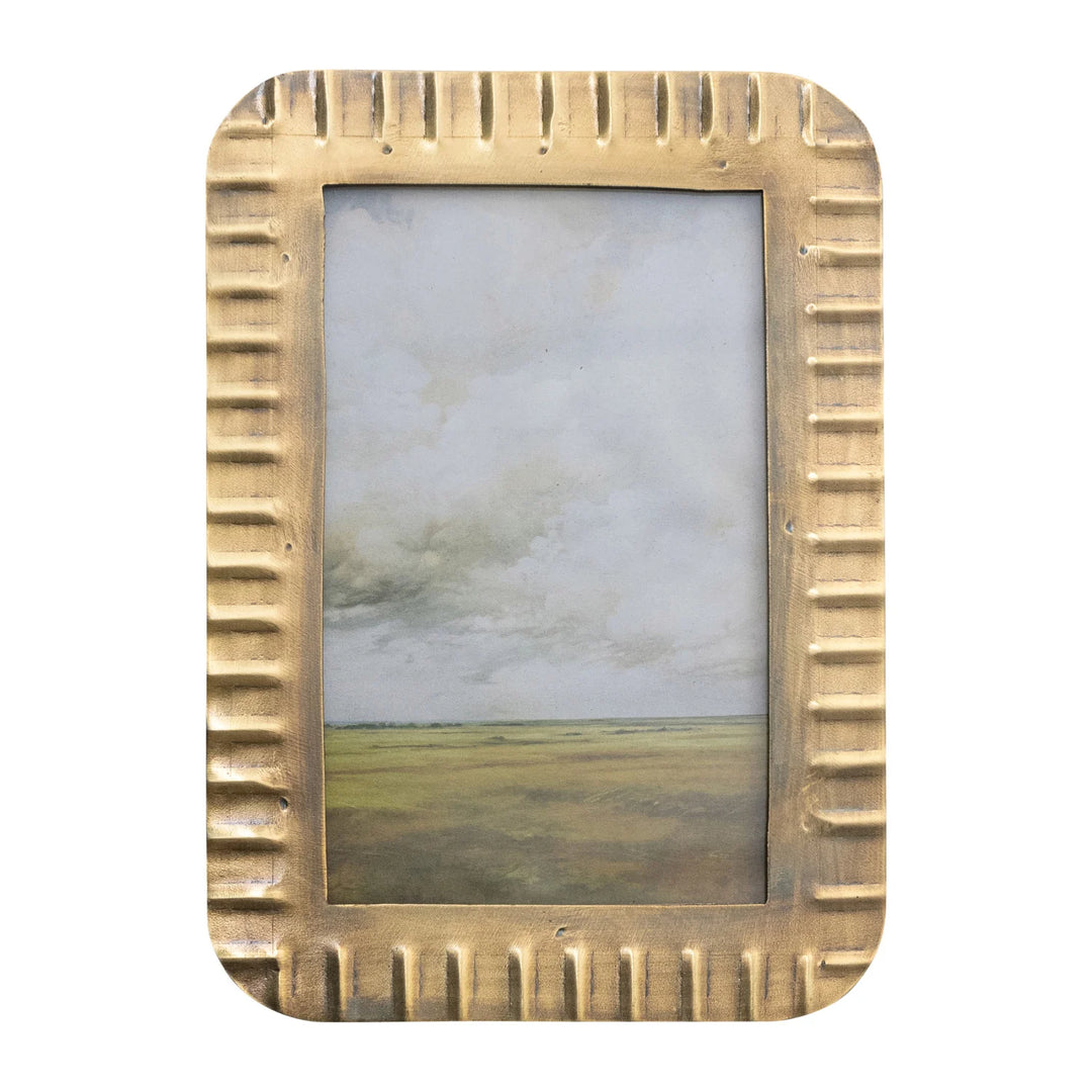A metal photo frame with a bold ribbed texture and warm antique brass finish, shown with a landscape painting inside.