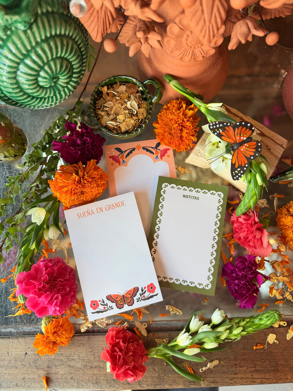 Two decorative notepads with butterfly illustrations are arranged on a rustic wooden surface surrounded by colorful flowers, dried petals, and terracotta pottery.