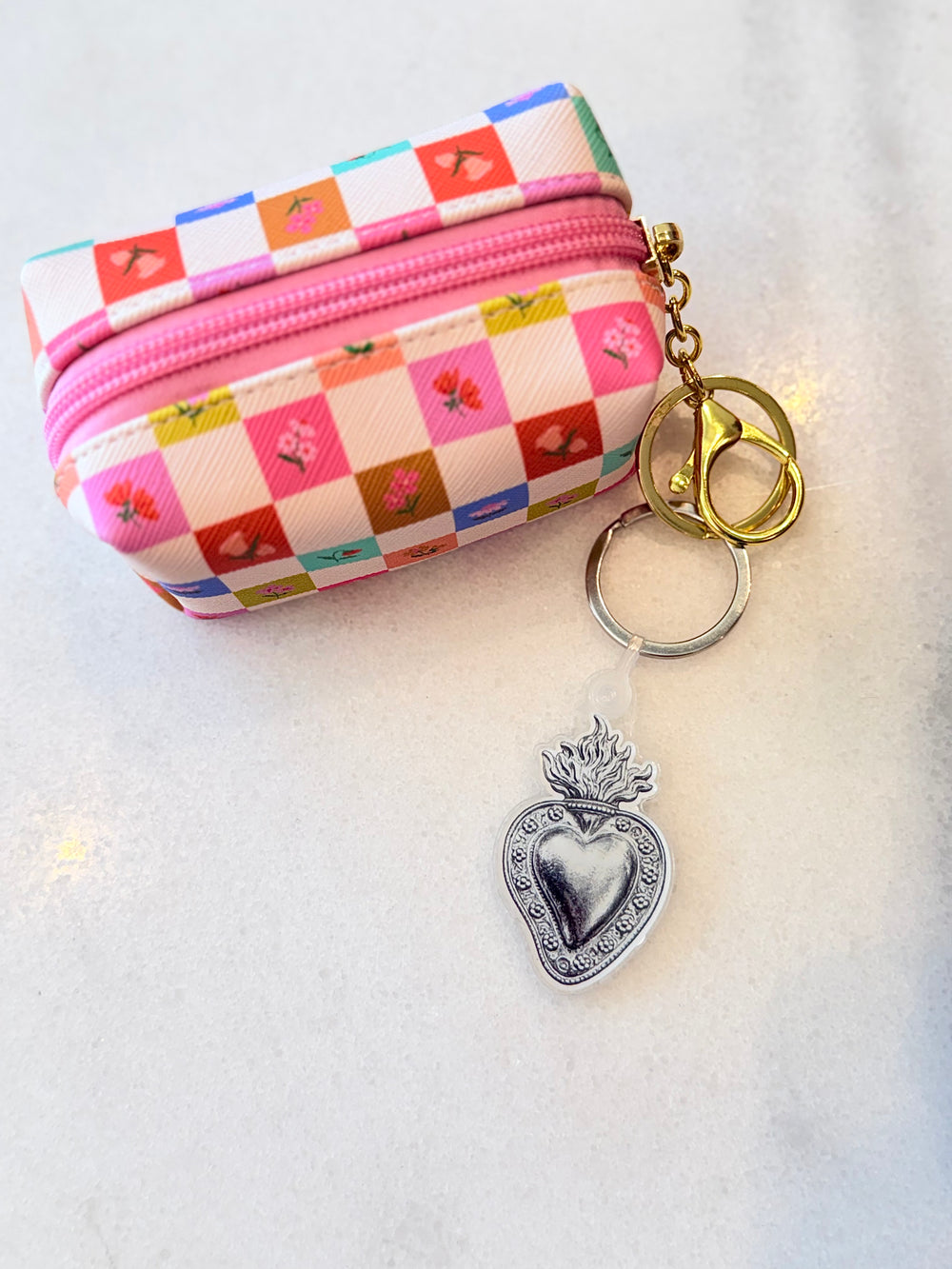A small zippered pouch with a colorful patchwork pattern of pink, red, orange, and blue squares, some featuring floral motifs. The pouch has a pink zipper and is shown with a keychain attachment featuring gold and silver rings and a silver Sacred Heart charm, all resting on a light textured surface.