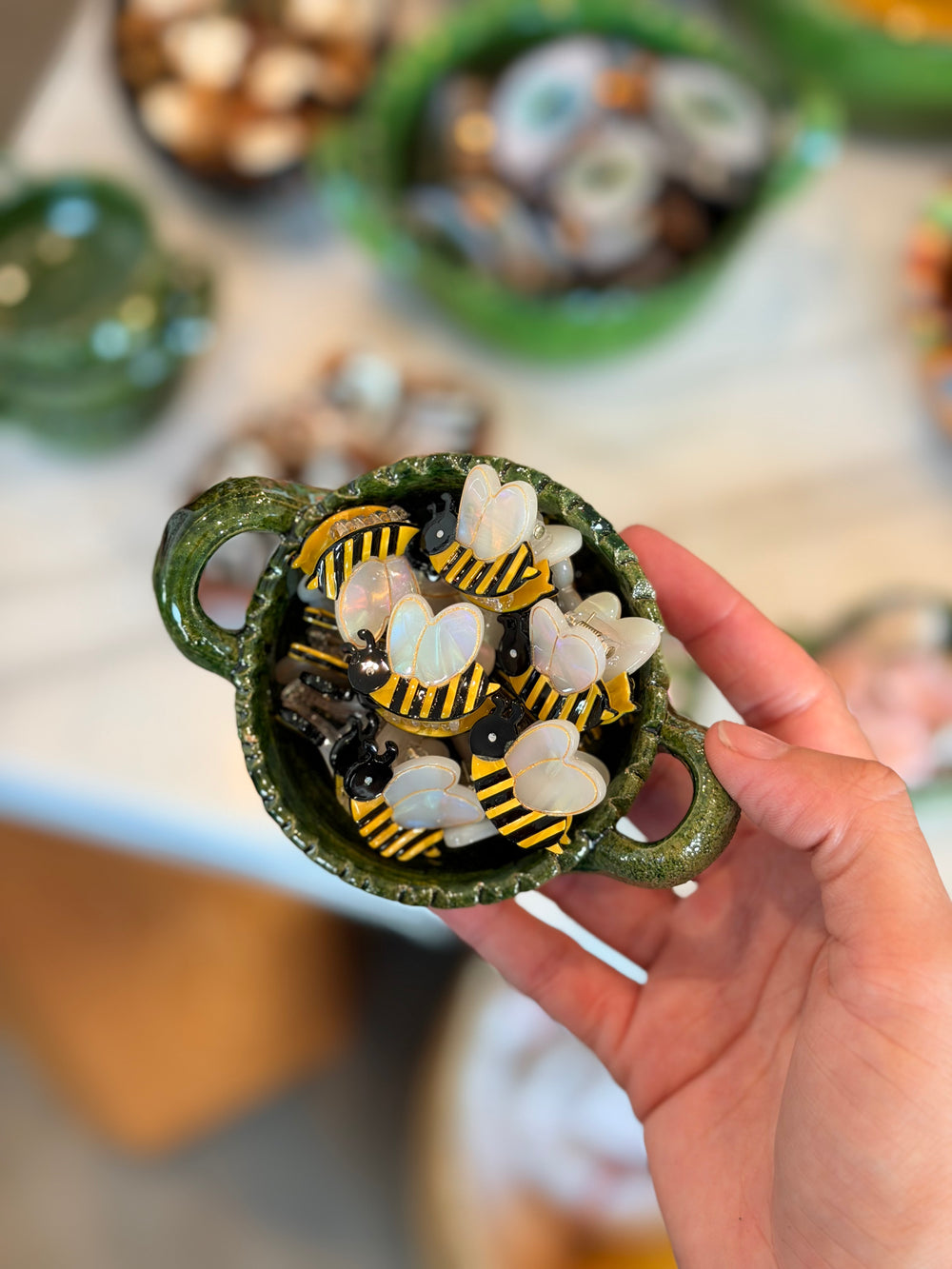 A hand holds a small green ceramic bowl filled with mini bumblebee hair clips. The figurines have white wings, yellow and black striped bodies.