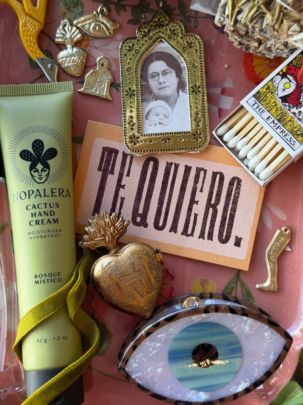 A styled flat-lay featuring a tube of Nopalera Cactus Hand Cream next to a pink card reading 'TE QUIERO.' with a gold heart-shaped locket, a gold-framed portrait photo, a matchbox, and other decorative objects on a patterned pink surface.