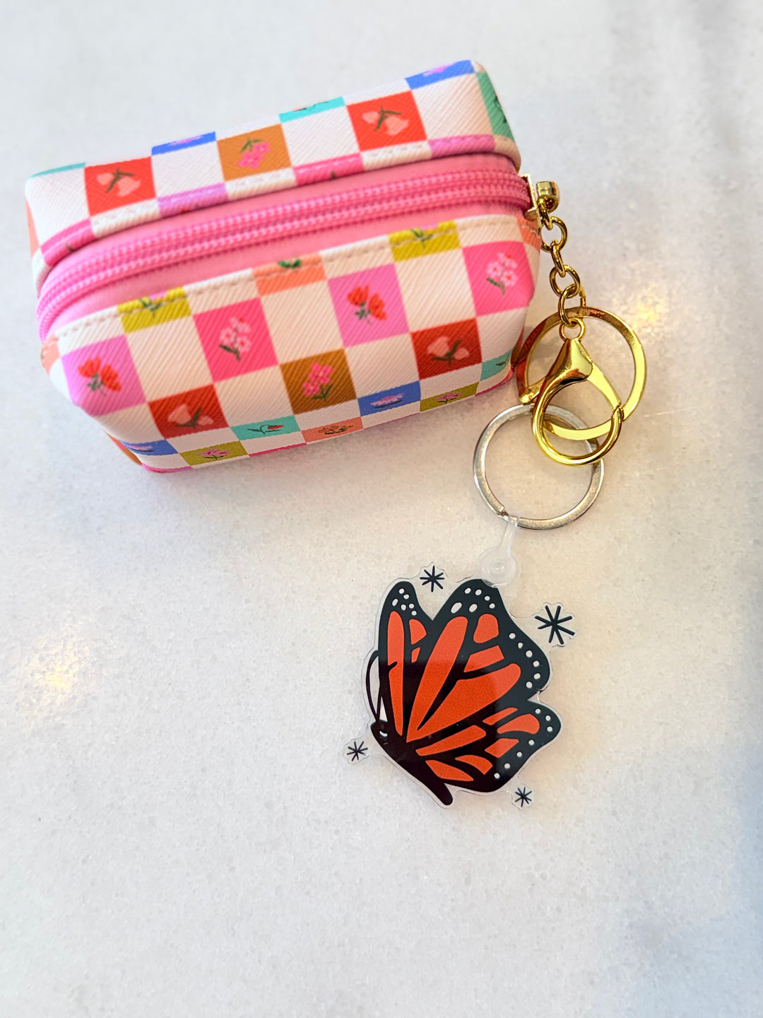 A small zippered coin purse with a colorful checkered pattern and floral print, shown with a monarch butterfly-shaped keychain on gold and silver rings on a light surface.