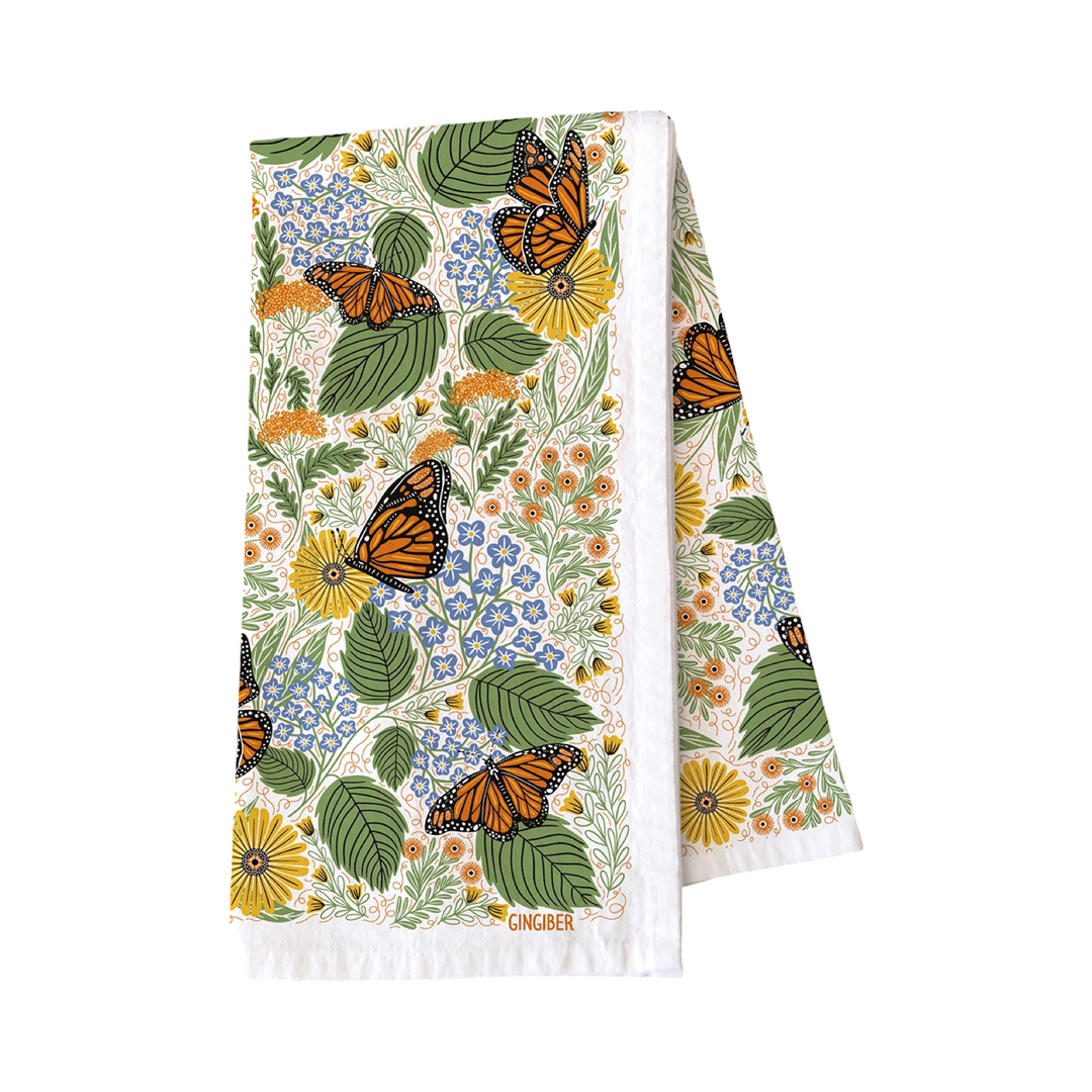 White folded kitchen towel features illustration of lush mix of flowers, greenery, and striking monarchs.