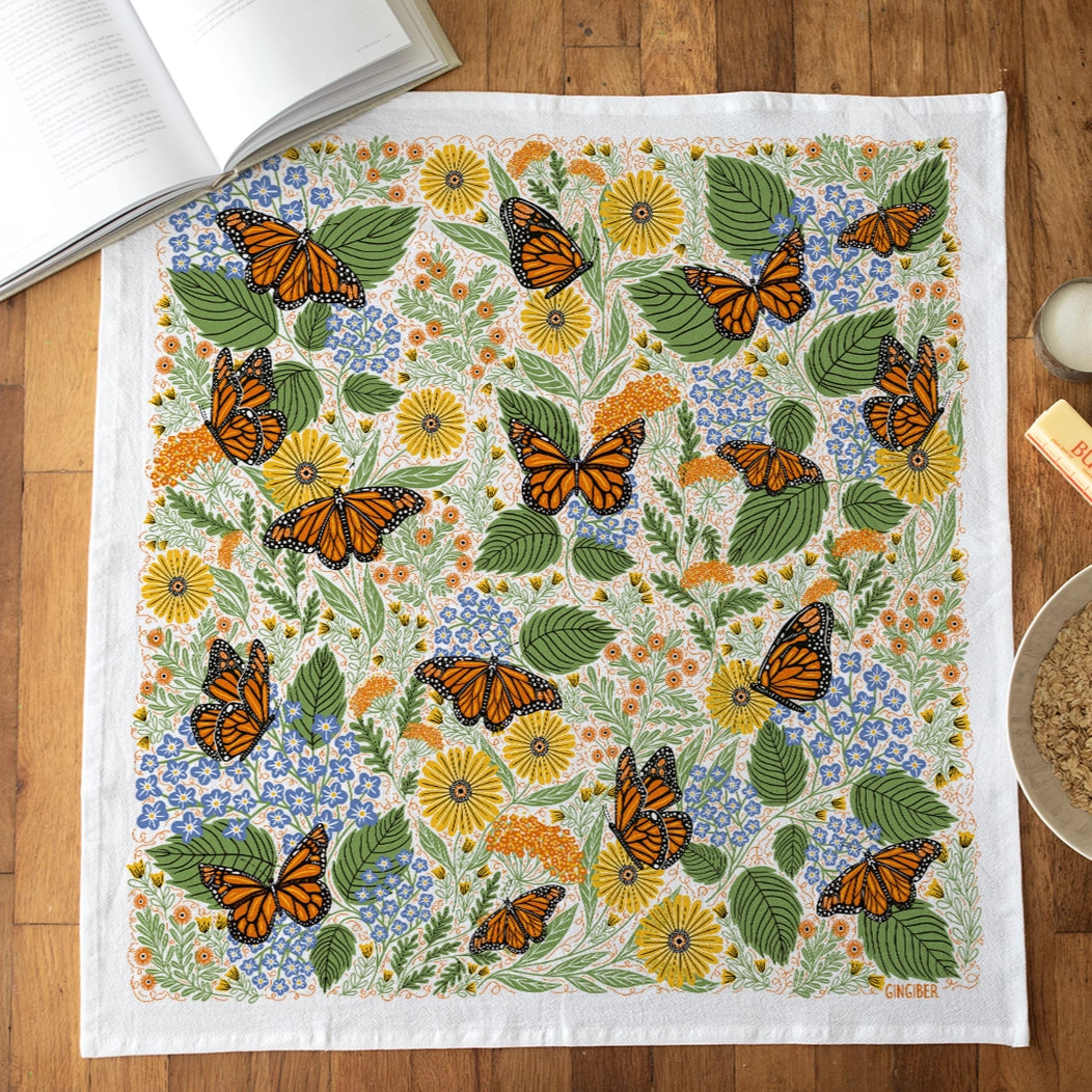 Flat towel on a wooden top features a  lush mix of flowers, greenery, and striking monarchs.