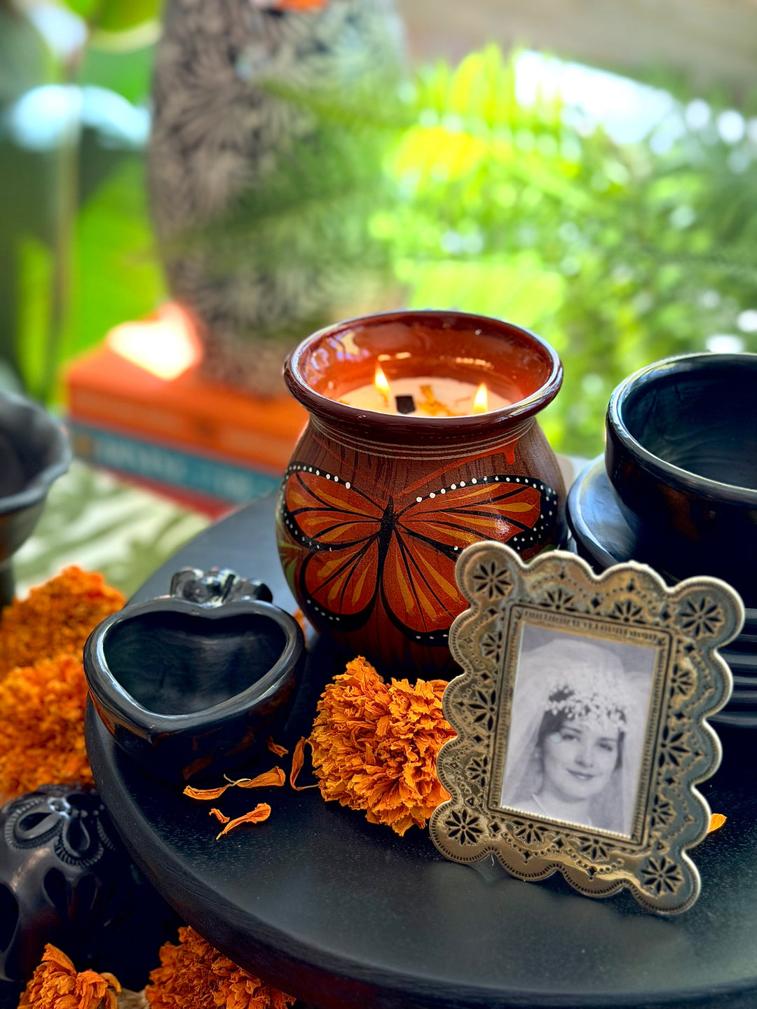 A decorative ceramic candle holder with a butterfly design, set on a black tray alongside orange marigold flowers, a small heart-shaped black bowl, and an ornate silver picture frame holding a black-and-white portrait, with a blurred green outdoor background.