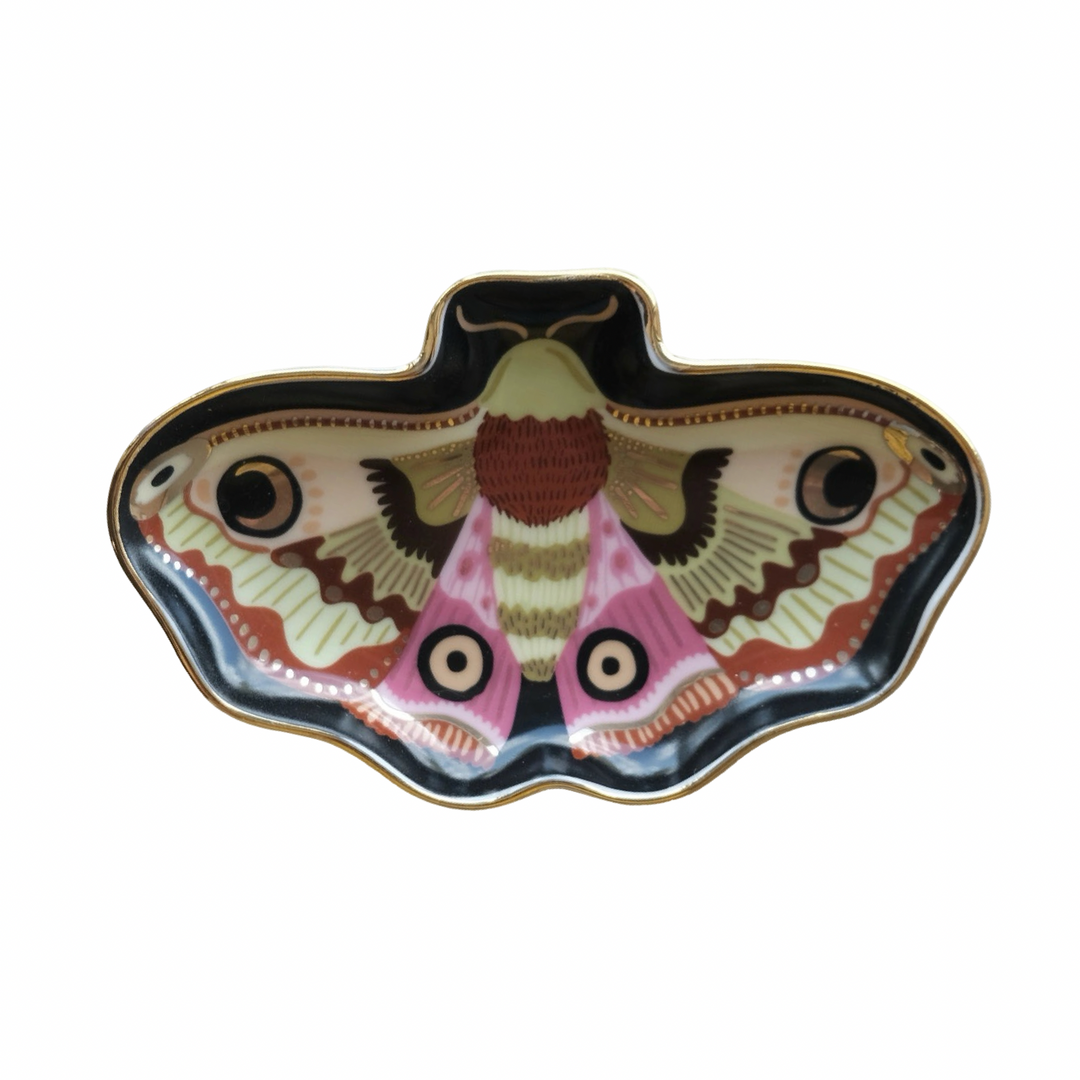 A moth shaped porcelain trinket dish with a detailed moon moth illustration in pink, cream, brown, and black tones with a gold rim, shown on a marble surface.