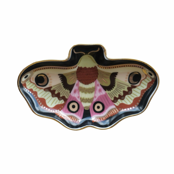 A moth shaped porcelain trinket dish with a detailed moon moth illustration in pink, cream, brown, and black tones with a gold rim, shown on a marble surface.