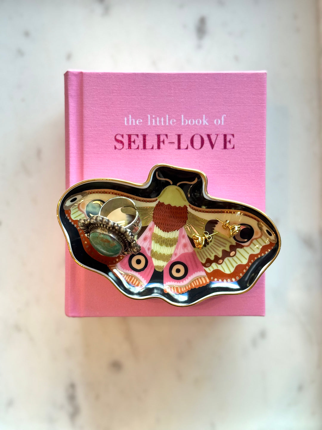 Pink book titled 'the little book of SELF-LOVE' with a moth design on a marble surface