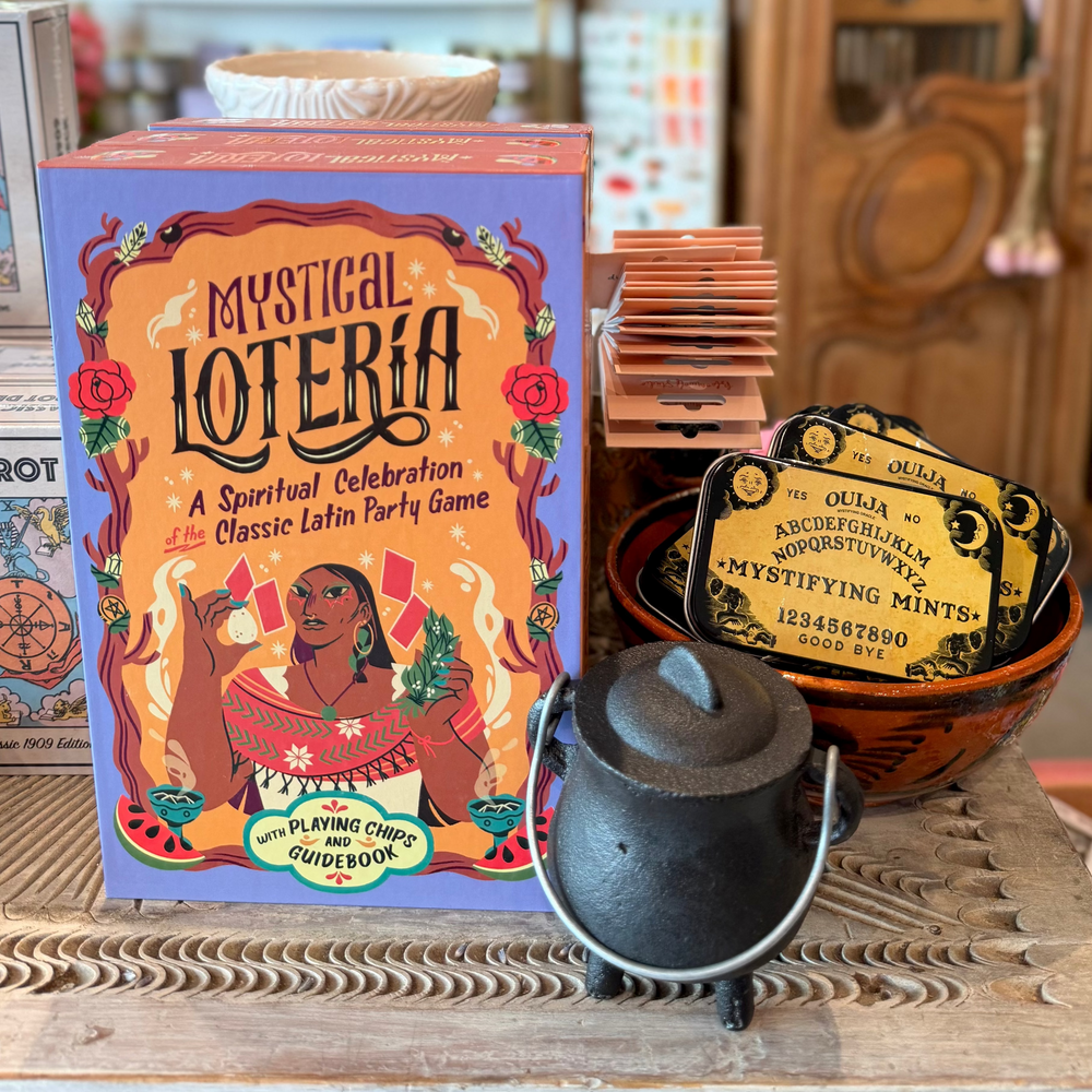 Mystical Loteria game box displayed on a wooden table in a shop setting, alongside a small black cauldron, a bowl of mystical mints, and other themed items.