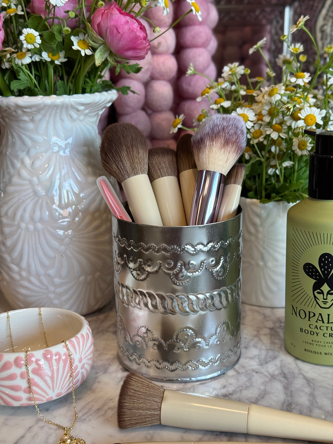 Makeup brushes in a decorative holder with flowers and a bottle of Nopal body cream on a marble surface.