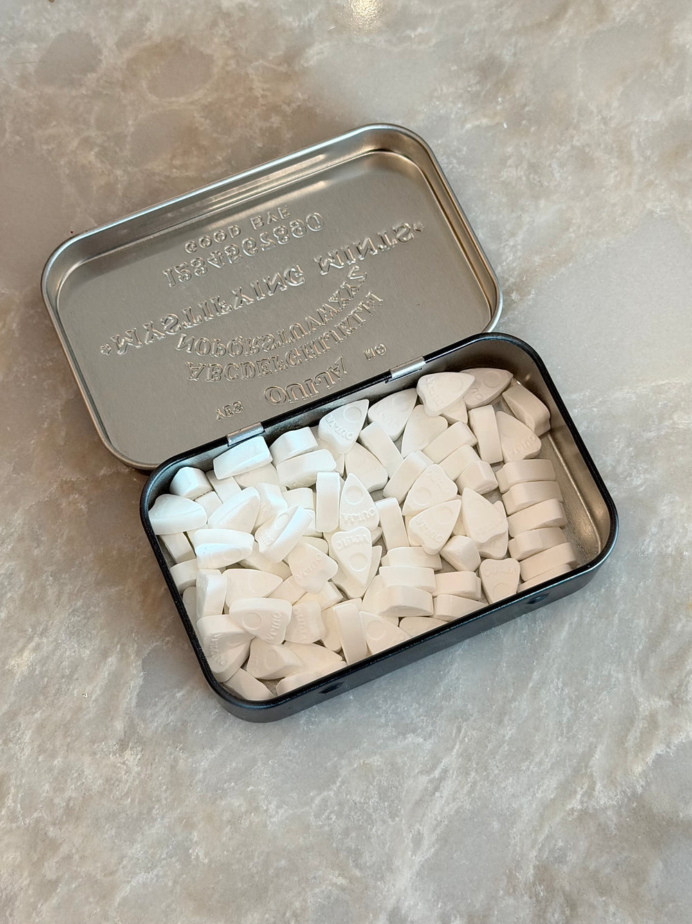 An open silver metal tin filled with many small white triangular pieces, placed on a light beige stone-like surface.