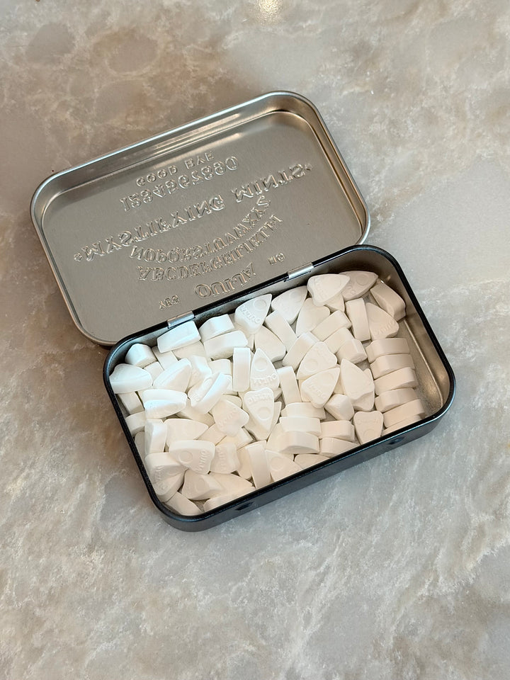 An open silver metal tin filled with many small white triangular pieces, placed on a light beige stone-like surface.