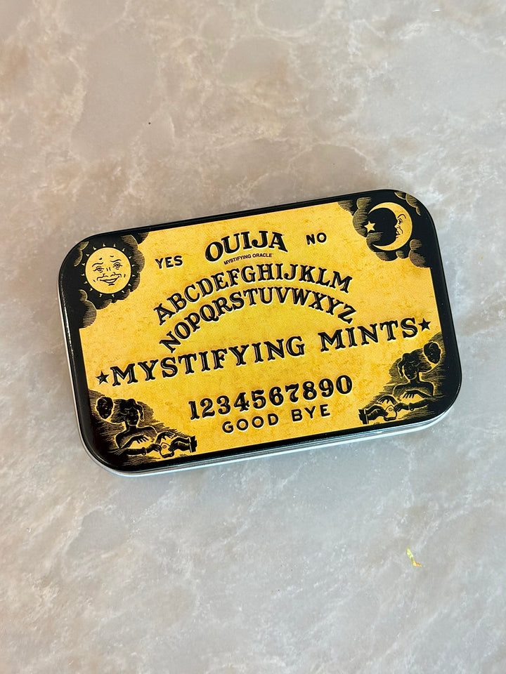 A rectangular tin of 'Ouija Mystifying Mints' with a yellow and black gothic Ouija board design, resting on a light beige marble surface.