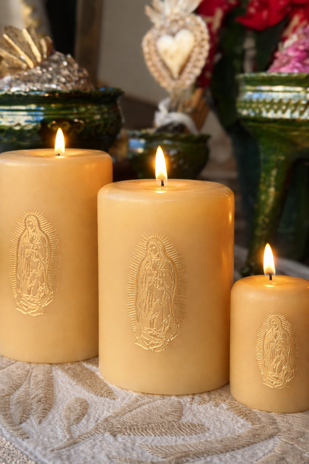 Three lit ivory pillar candles in assorted sizes lined up in a graduated row feature gold embossed Our Lady de Guadalupe design. They sit on a ivory and beige Otomi textile with home decor in the background.