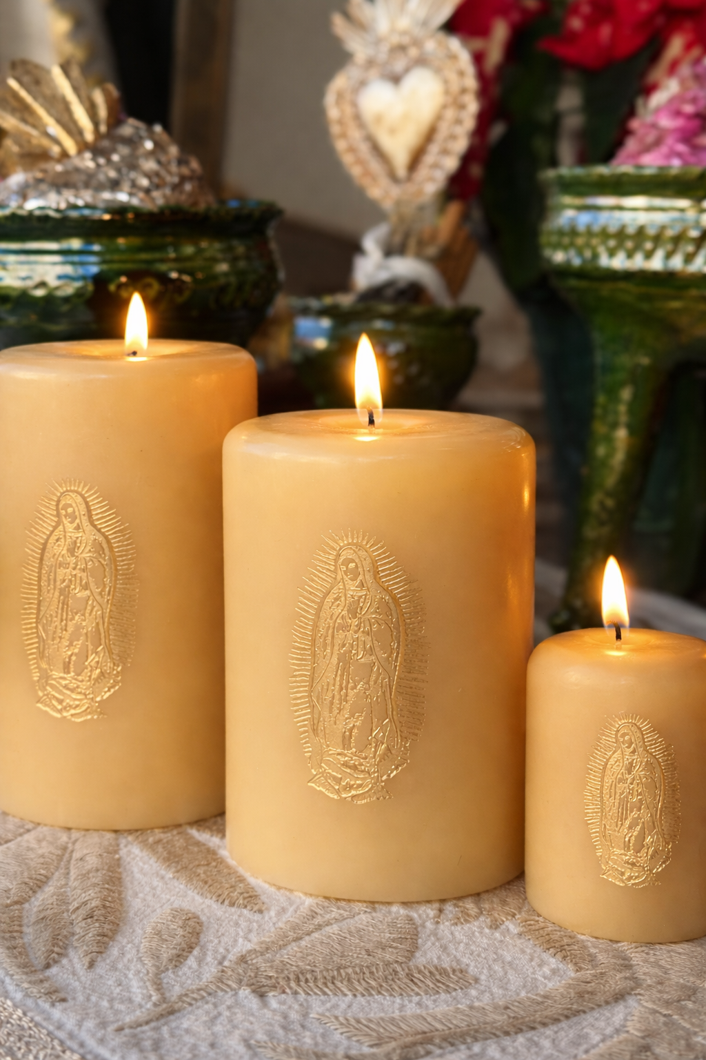 Three lit ivory pillar candles in assorted sizes lined up in a graduated row feature gold embossed Our Lady de Guadalupe design. They sit on a ivory and beige Otomi textile with home decor in the background.