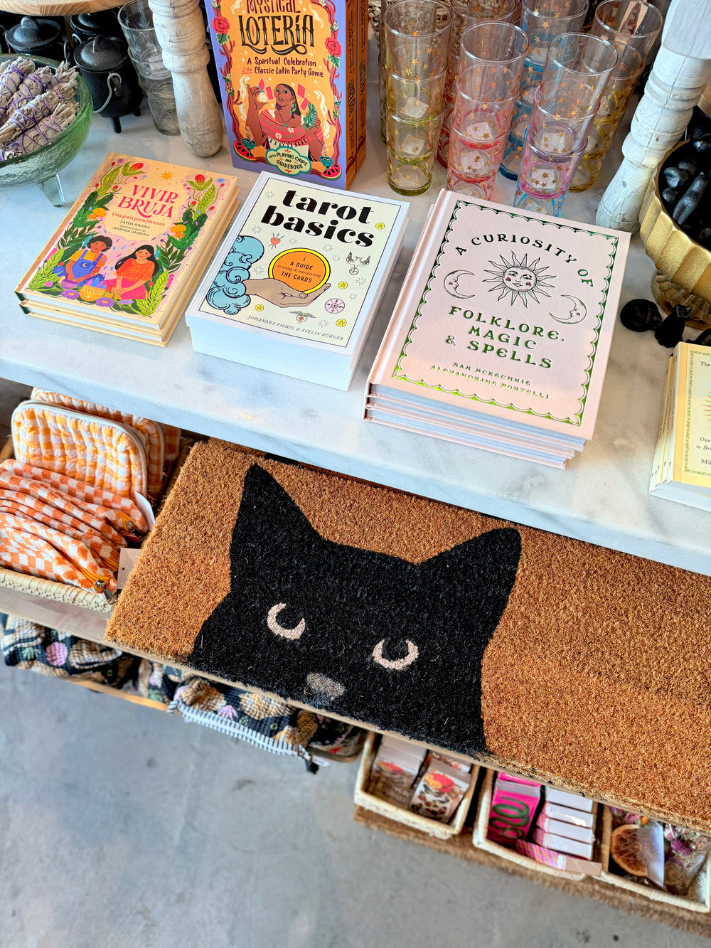 A brown coir doormat with a large black cat face and small white crescent moon eyes, displayed on a store shelf alongside books and other home goods.