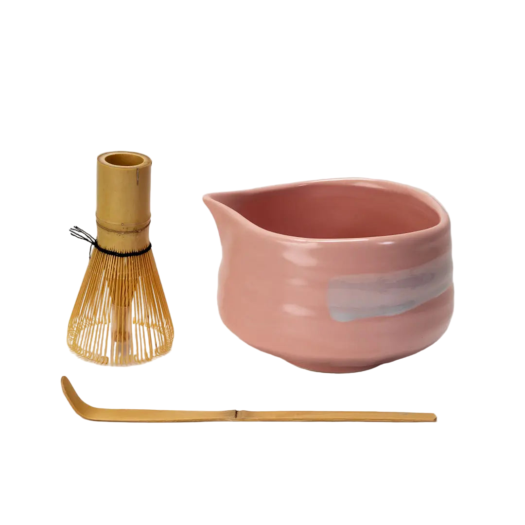 Side view of a pink matcha bowl, whisk and bamboo spoon all on a white background.