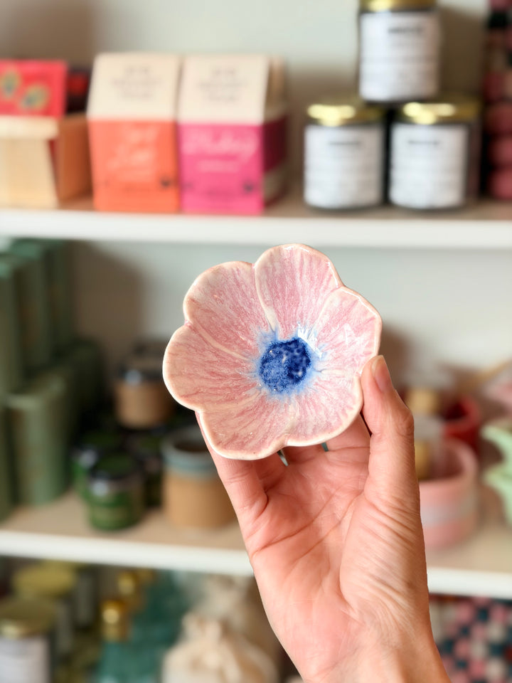 Hand holding a pink ceramic flower bowl with blue center against a blurred background of shelves with products.