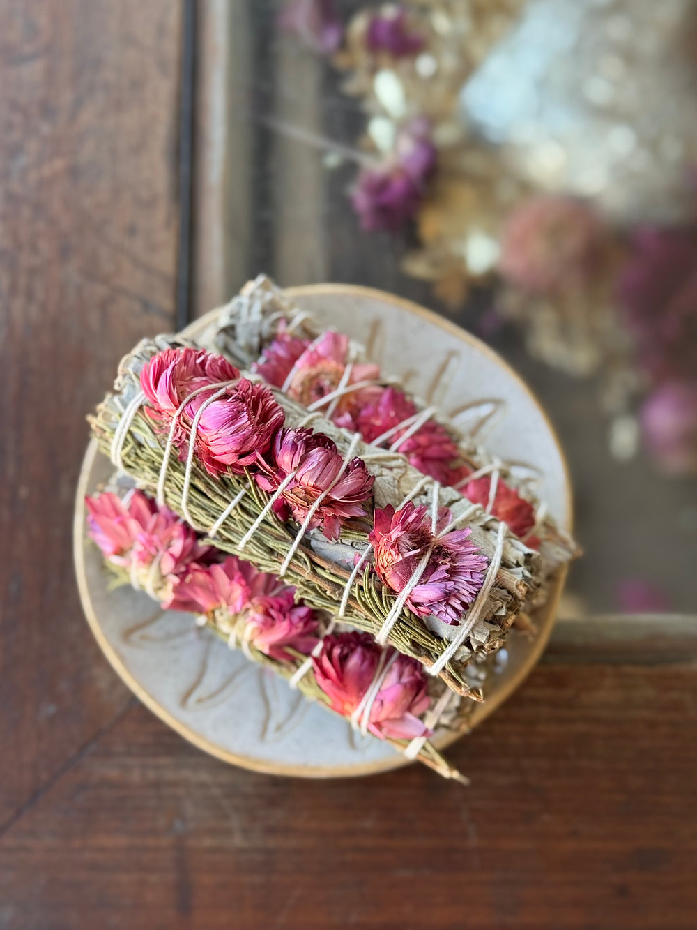 Smudge sticks wrapped in sage and rosemary, decorated with bright pink sunflower petals, tied together with natural twine are stacked in a dish on a wood and glass tabletop.