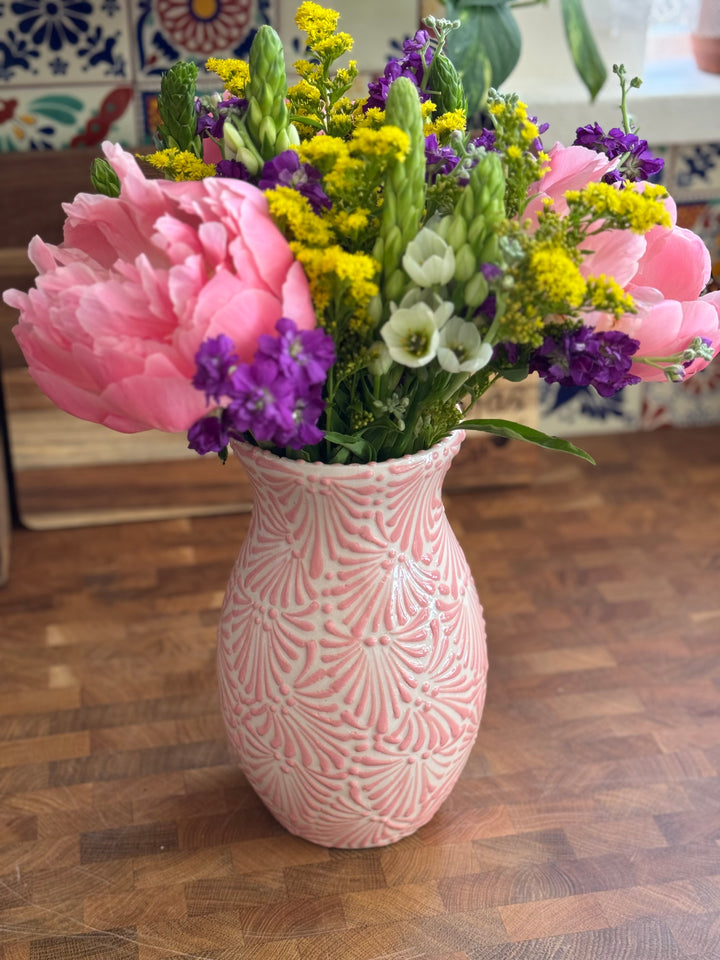 Decorative pink talavera vase with a colorful floral arrangement on a wooden cutting board surface.