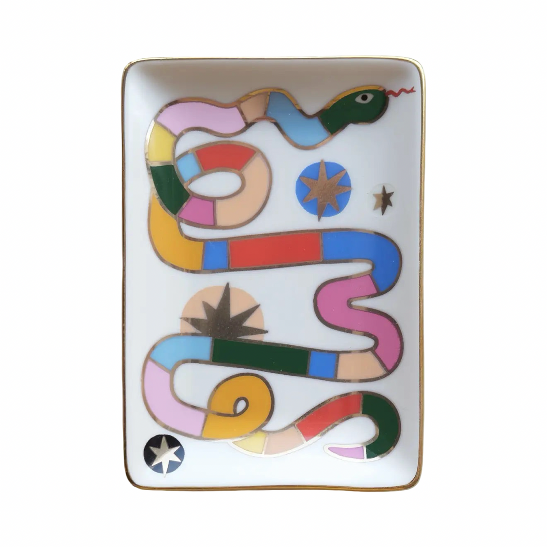A rectangular porcelain trinket dish featuring a colorful illustrated snake in multicolor patchwork segments with gold star accents and a gold rim, shown on a light surface.