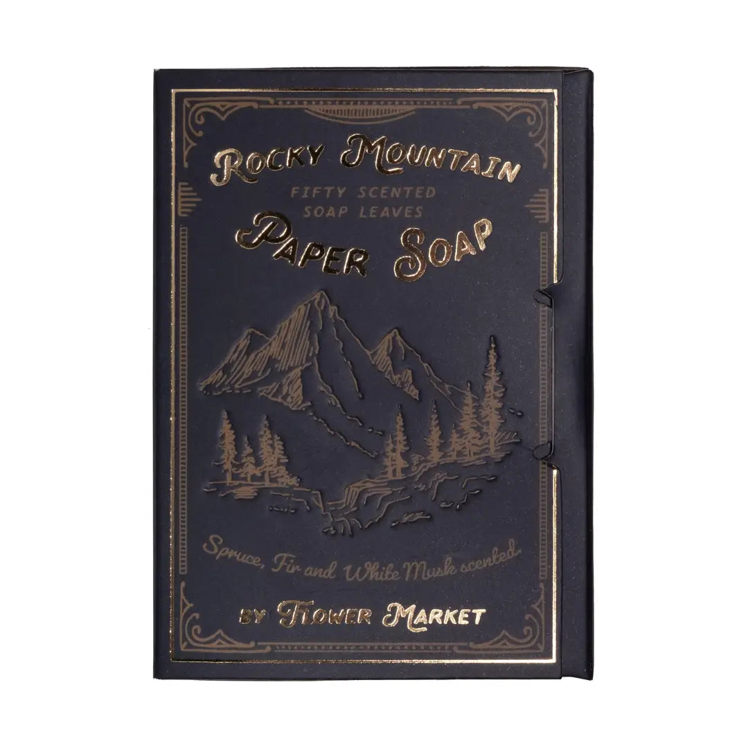 A dark navy book style package of Rocky Mountain paper soap leaves by Flower Market, featuring gold foil mountain and pine tree illustrations, scented with spruce, fir, and white musk, containing 50 soap leaves.