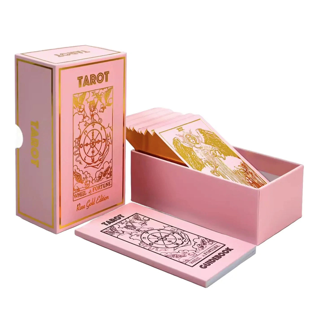 Pink rose-gold tarot deck displayed with cards partly fanned in an open box and a matching guidebook beside it.
