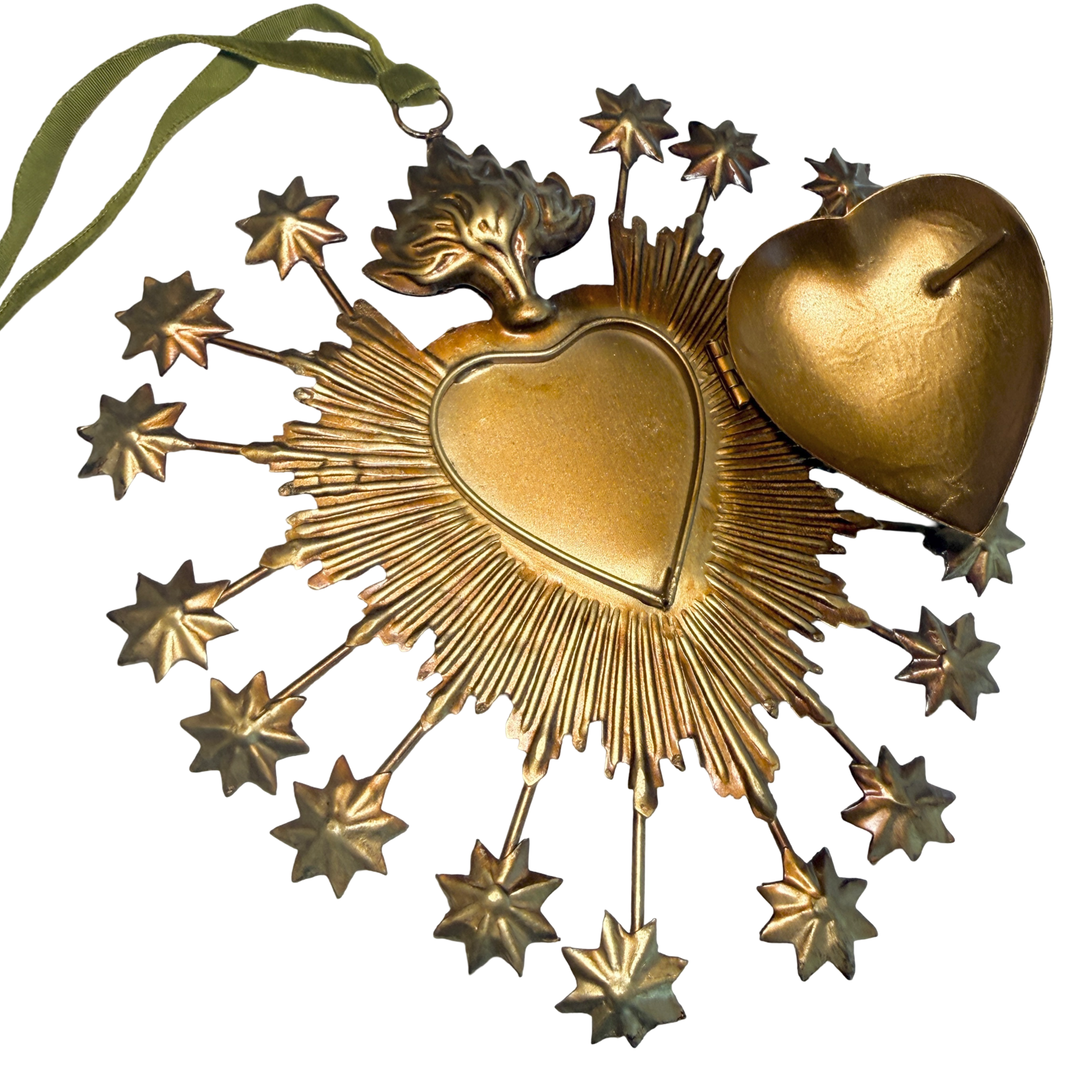 A gilded metal sacred heart locket box shown open, revealing the hollow interior of the heart-shaped vessel, with a radiating sunburst, star-tipped rays, and flame topper. Green velvet ribbon for hanging visible