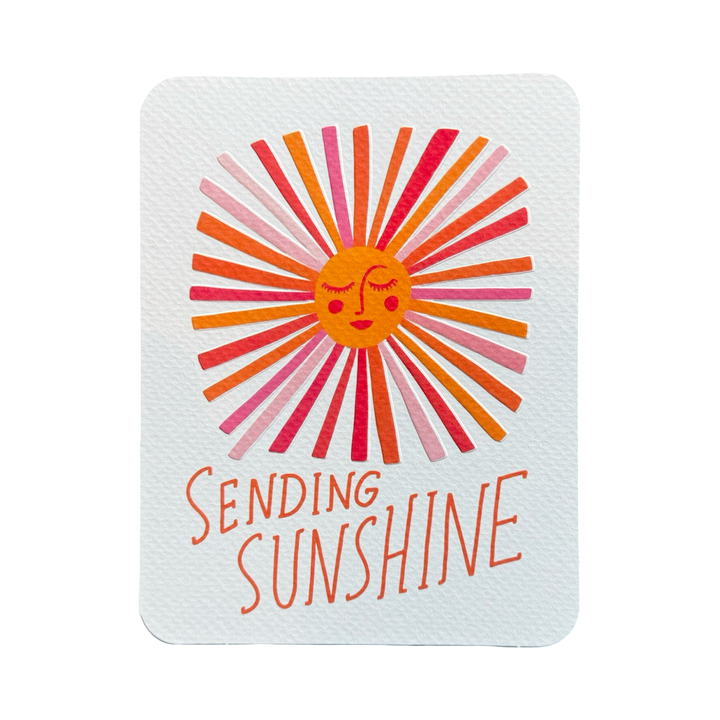 WHITE CARD WITH A SUN WITH RAYS IN VARIOUS SHADES OF ORANGE AND PINK WITH THE PHRASE SENDING SUNSHINE IN ORANGE LETTERING.