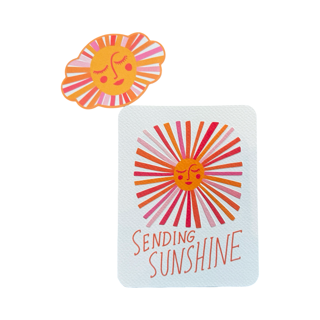 WHITE CARD WITH A SUN WITH RAYS IN VARIOUS SHADES OF ORANGE AND PINK WITH THE PHRASE SENDING SUNSHINE IN ORANGE LETTERING AND A MATCHING SUN STICKER.
