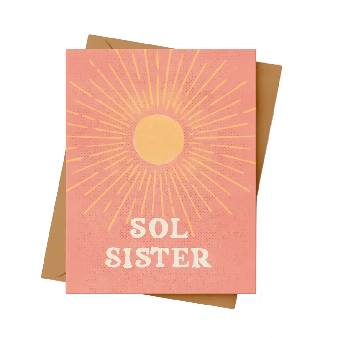 A greeting card with a warm coral background featuring a golden sunburst radiating from a circular sun in the upper half, with the words 'Sol Sister' in bold cream retro lettering at the bottom. Card is shown on kraft colored envelope.