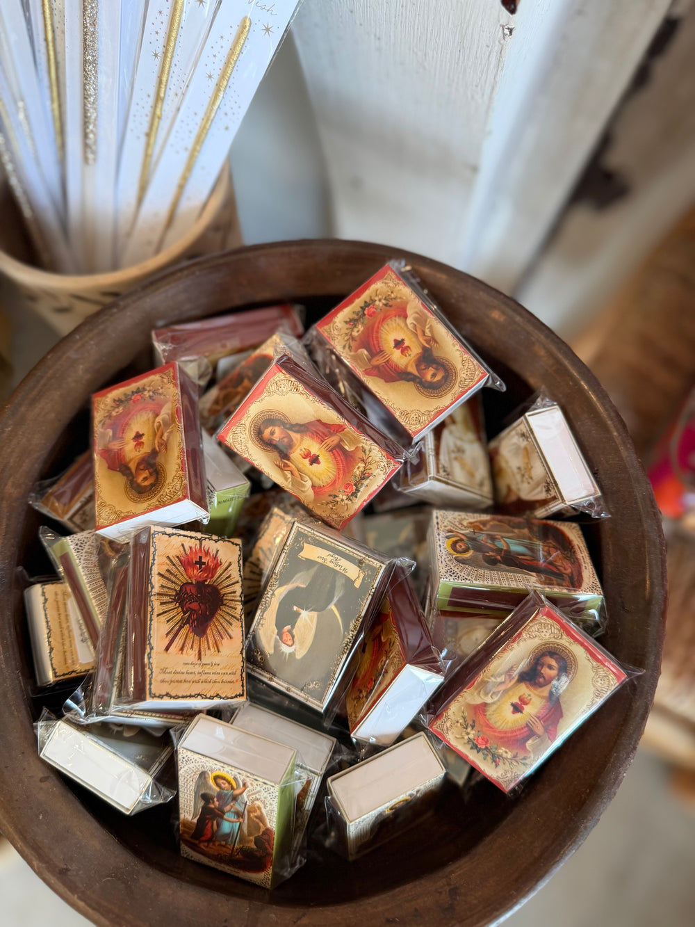 A wooden bowl filled with small packaged religious match boxes featuring colorful saint and icon illustrations, photographed inside Casa Palma Dulce