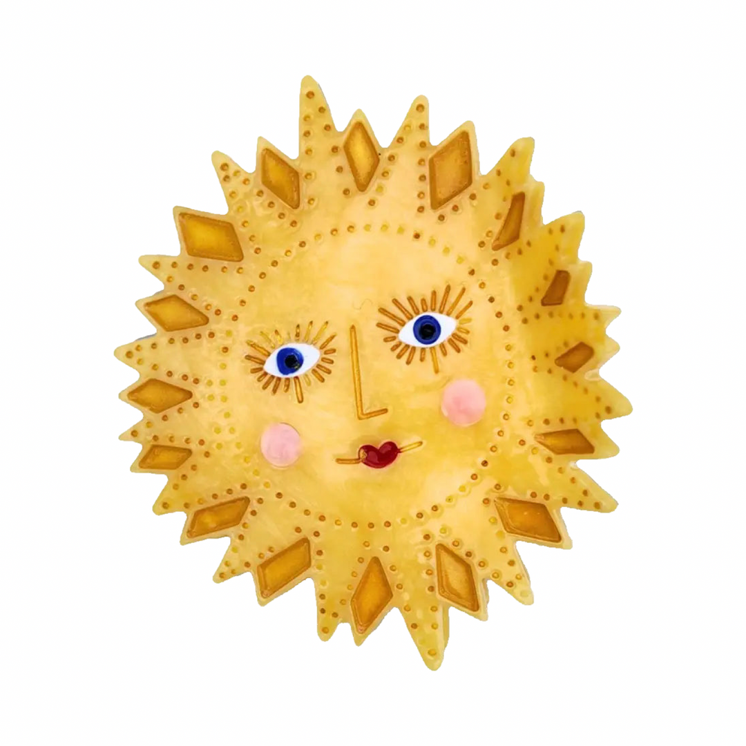 A yellow cellulose acetate sun-shaped claw hair clip with a hand-painted face featuring blue eyes, rosy cheeks, and a red heart mouth, with amber diamond ray detailing and dot accents. 