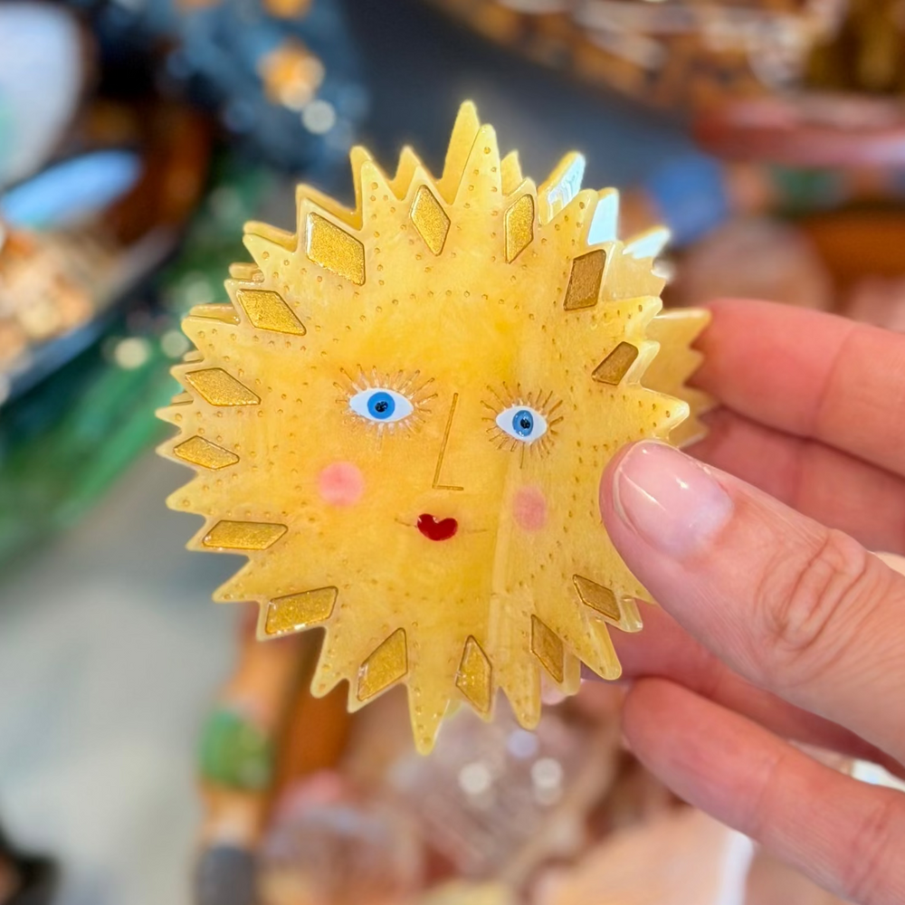 A yellow cellulose acetate sun-shaped claw hair clip with a hand-painted face featuring blue eyes, rosy cheeks, and a red heart mouth, with amber diamond ray detailing and dot accents. 