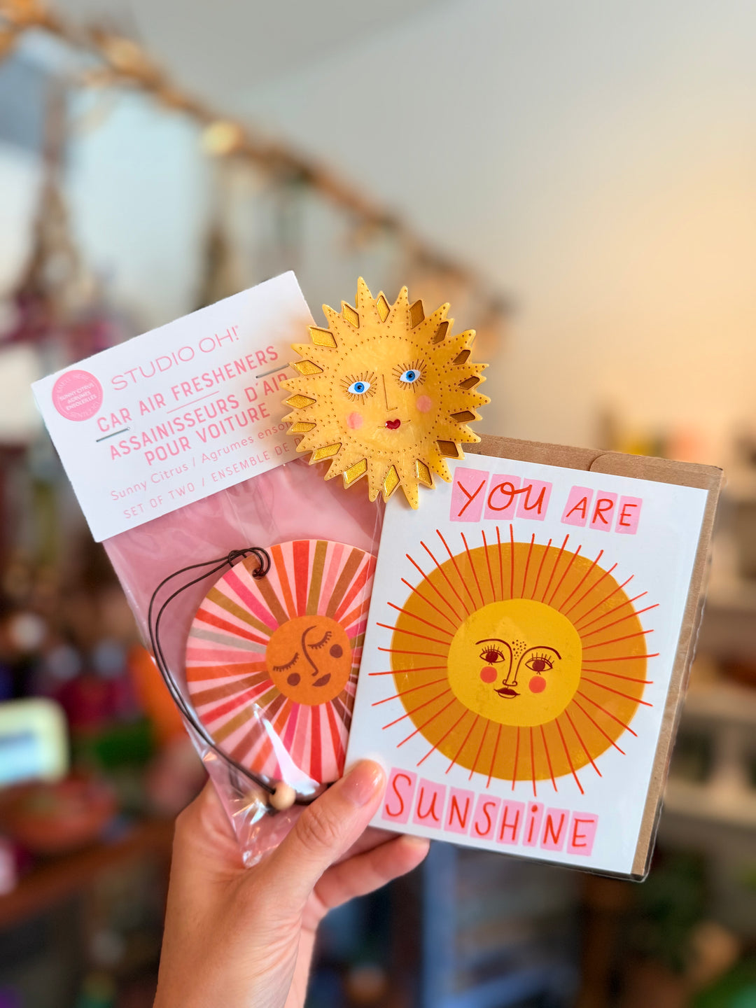 A hand holds a Studio Oh! car air freshener set and a greeting card. The set includes a pink and orange sun-themed cap air freshener in its packaging and a matching greeting card that reads 'YOU ARE SUNSHINE' with a smiling sun illustration.