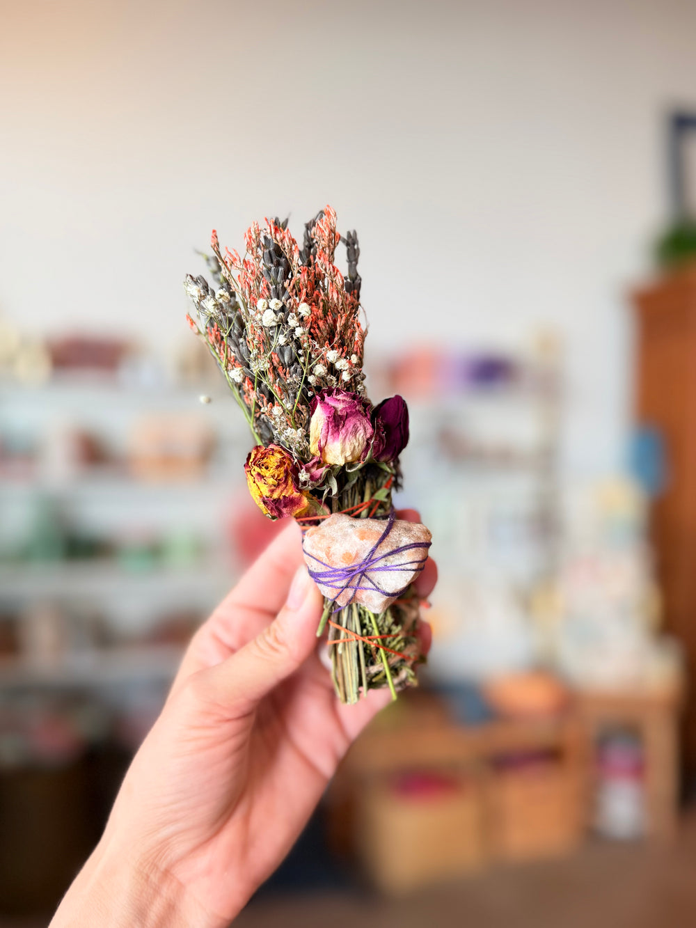 A hand holding a small dried flower bouquet with pink, purple, orange, and white botanicals tied with a purple ribbon, set inside of Casa Palma Dulce