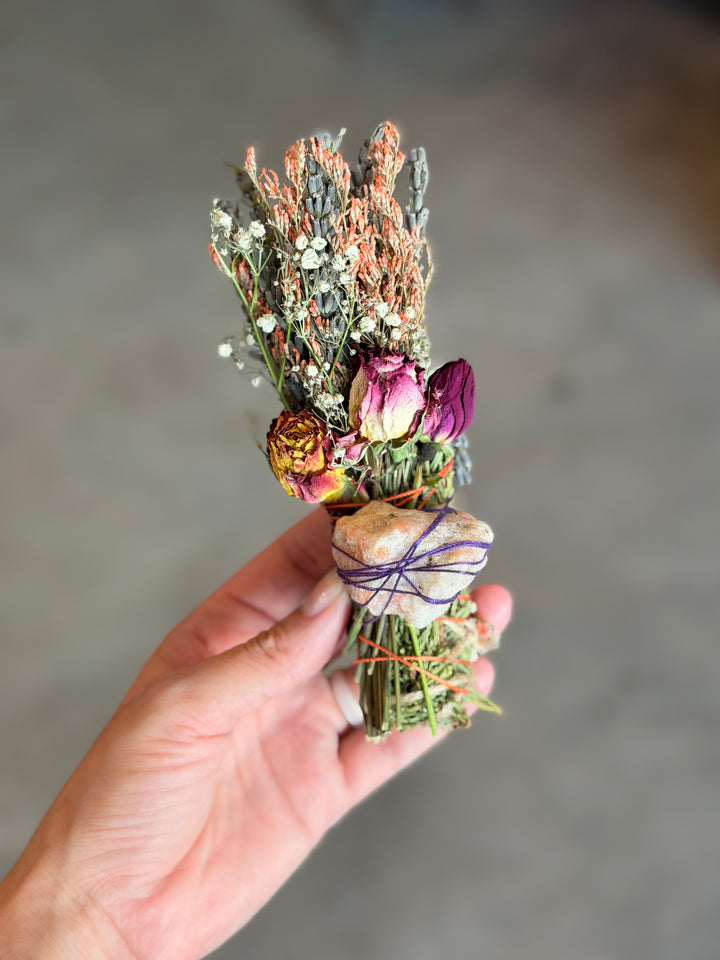 A hand holding a small dried flower bouquet with purple, pink, and orange blossoms tied with colorful string