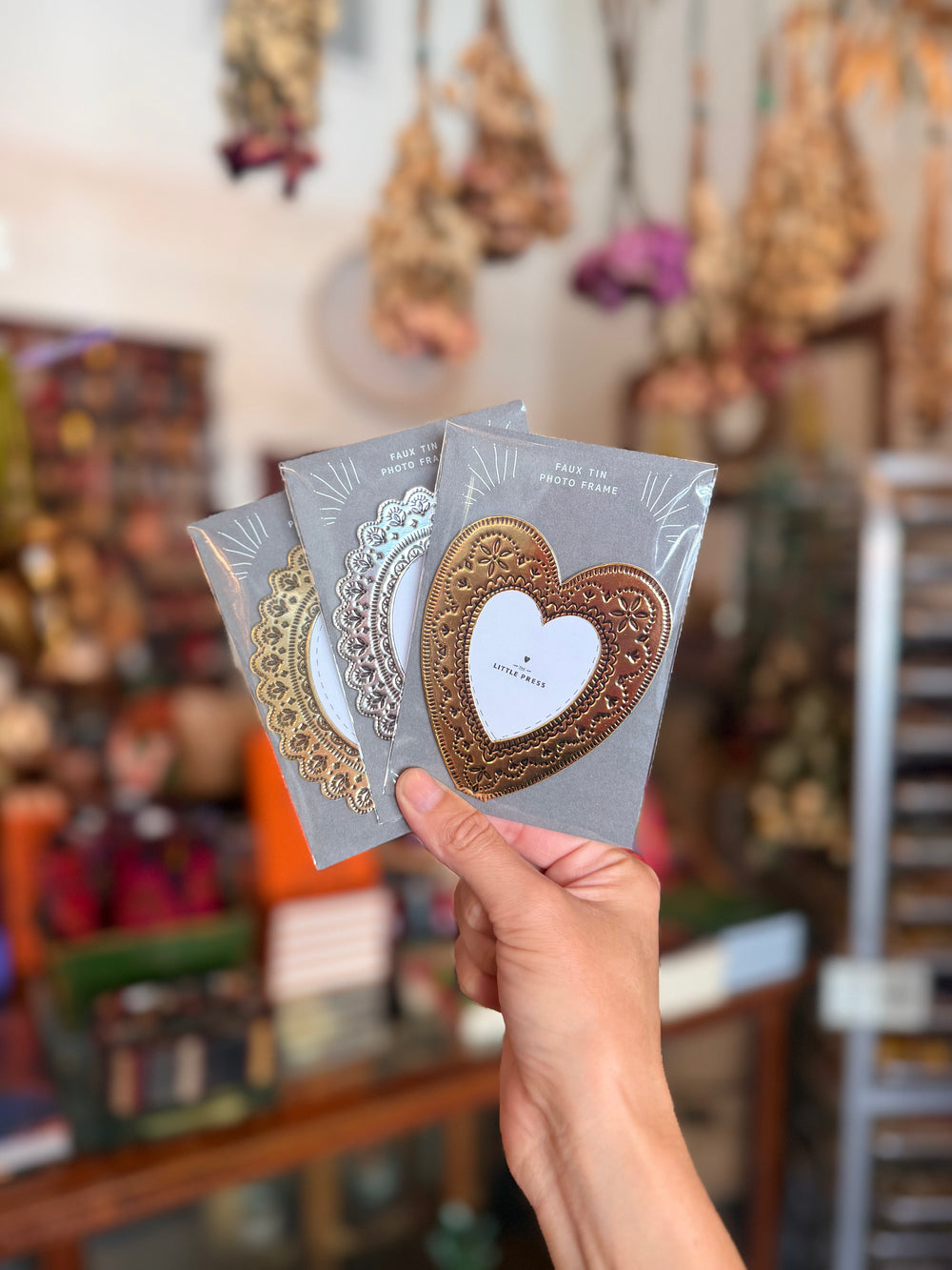 A hand holds three decorative metal heart-shaped frames in different gold, silver, and rose gold finishes, each mounted on a gray backing card. 