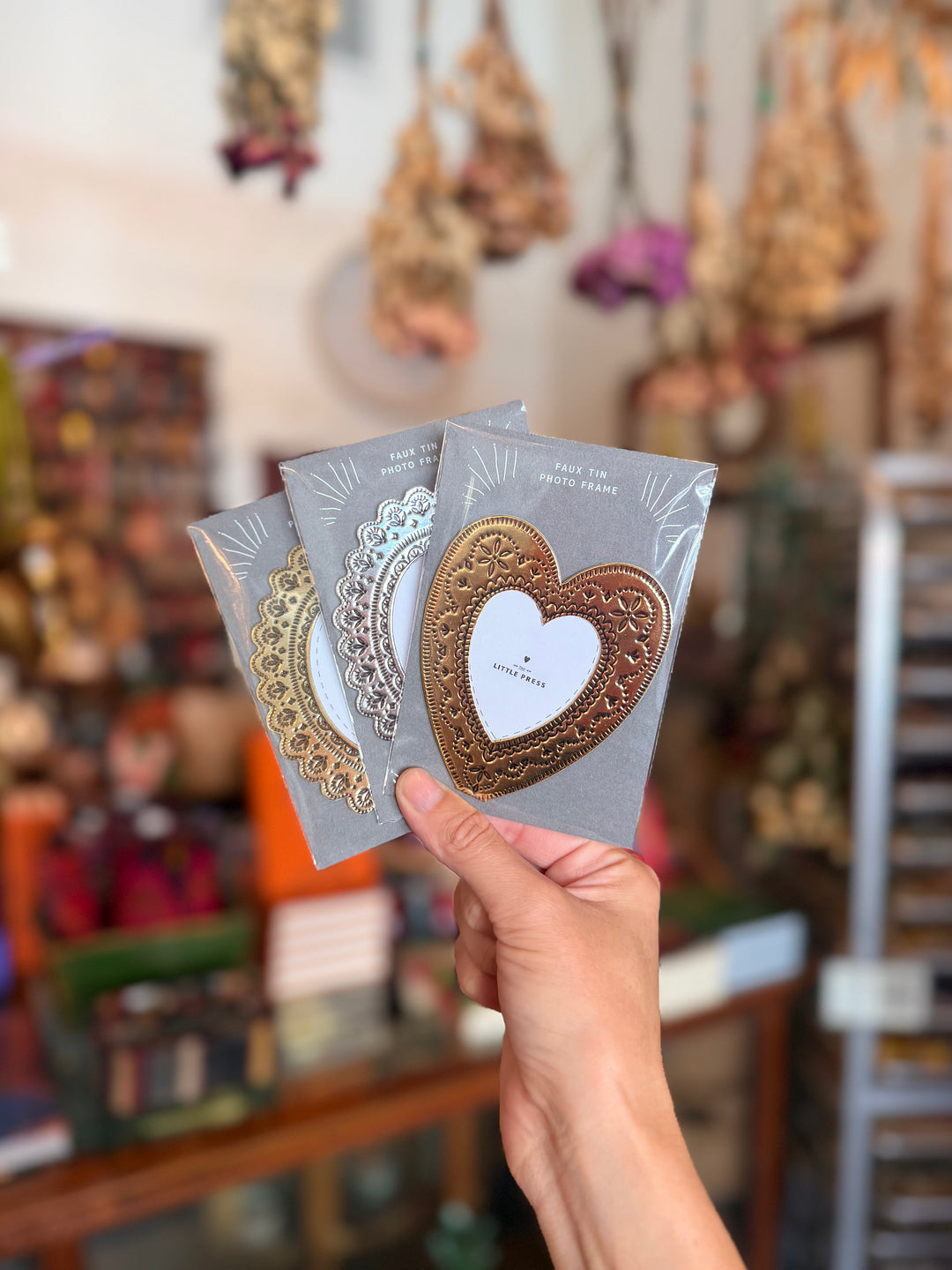 A hand holds three decorative metal heart-shaped frames in different gold, silver, and rose gold finishes, each mounted on a gray backing card. 