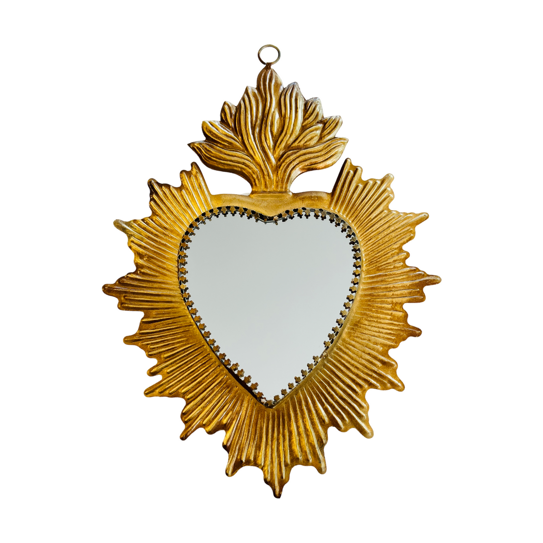 A gold-colored tin mirror with a heart-shaped reflective surface, surrounded by a floral-like design, hanging on a wall.