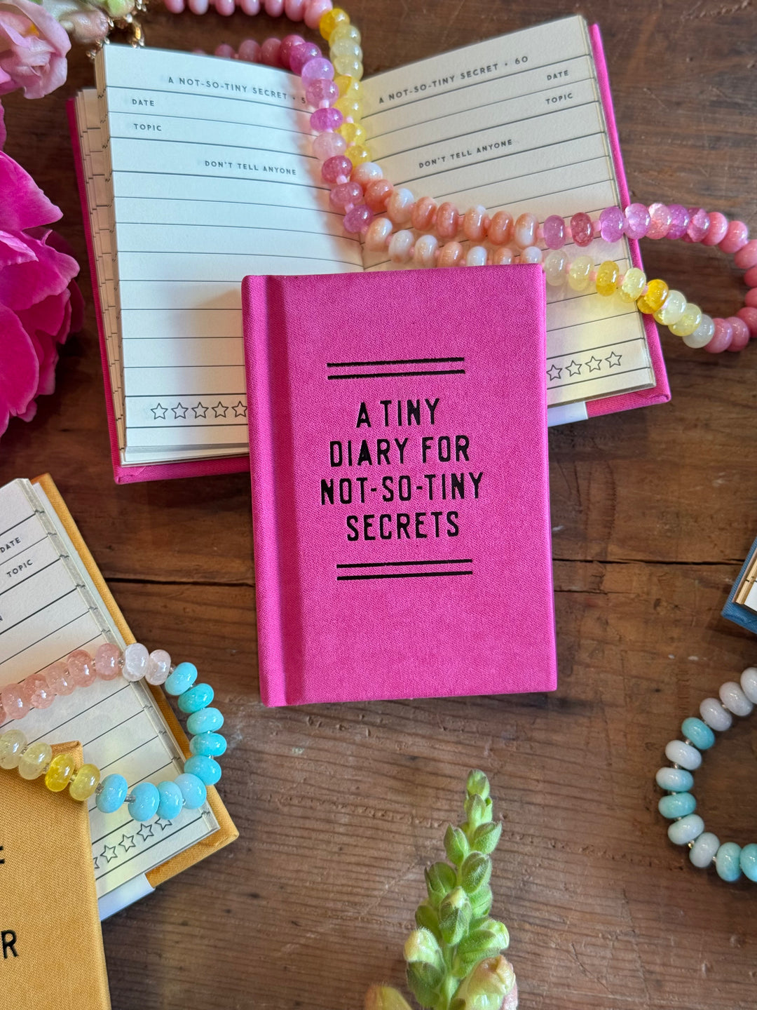 Pink diary titled 'A Tiny Diary for Not-So-Tiny Secrets' surrounded by colorful bracelets on a wooden surface.