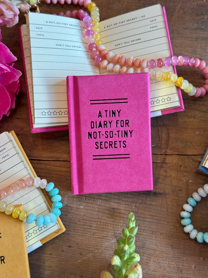 Pink diary titled 'A Tiny Diary for Not-So-Tiny Secrets' surrounded by colorful bracelets on a wooden surface.