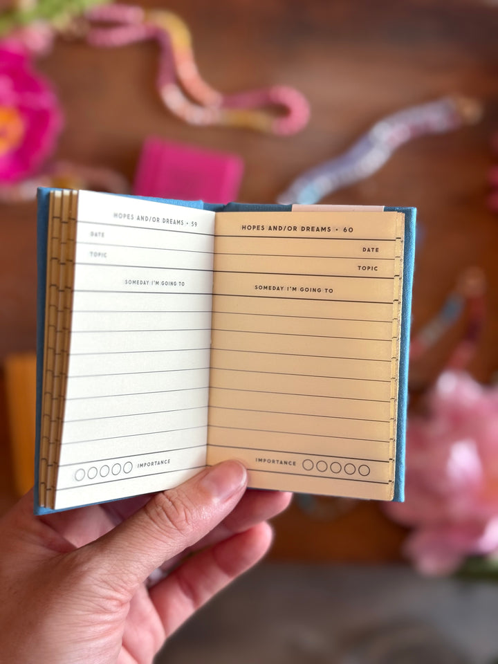 Hand holding a small notebook with lined pages and a blue border, against a blurred background with flowers.