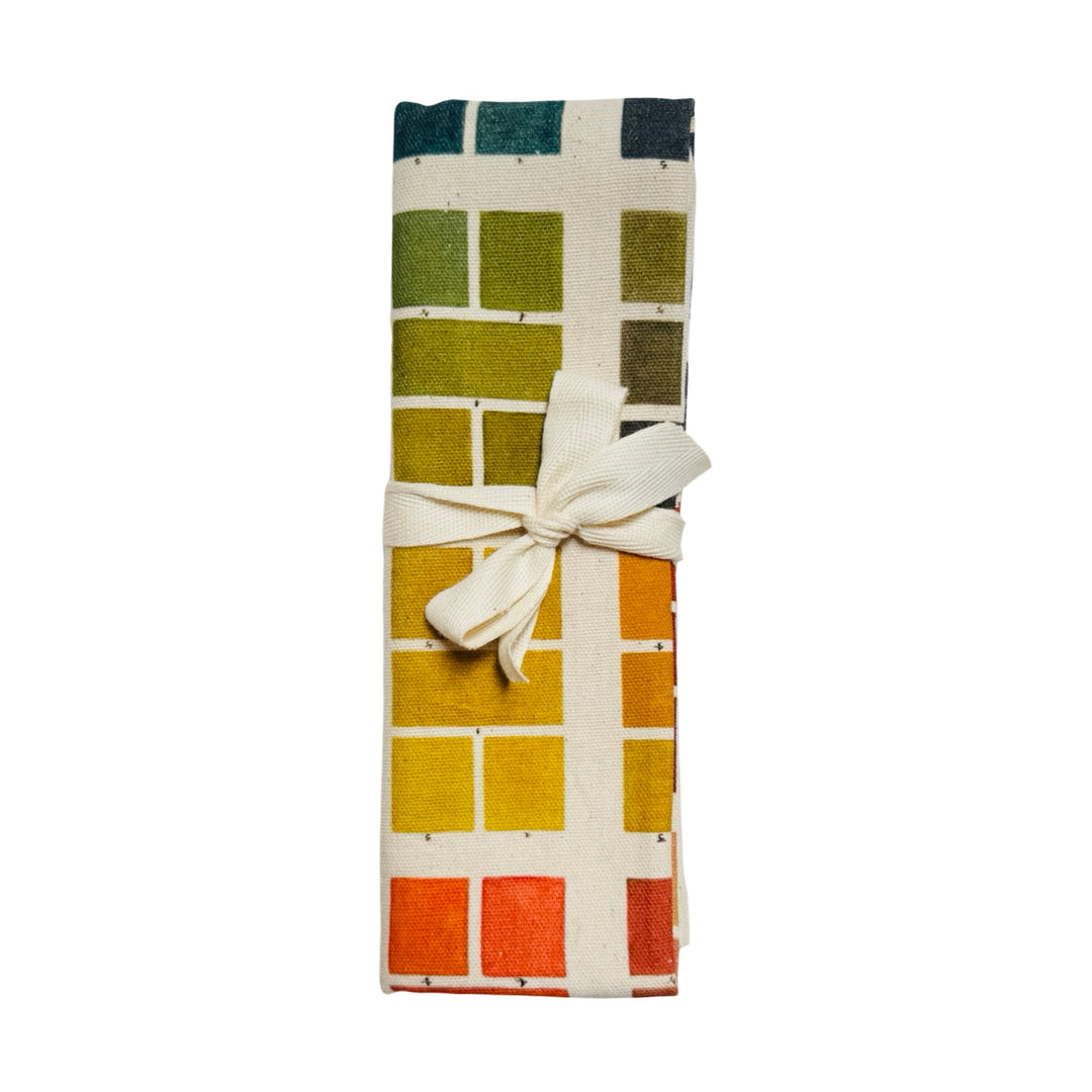 Tea towel featuring a grid of watercolor paint swatches in a range of colors with French text printed at the top folded in half and tied with a ribbon.