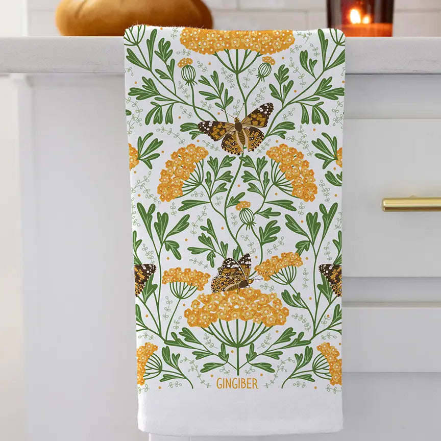 White cotton tea towel printed with a botanical pattern of golden yarrow flowers and painted lady butterflies, accented with green leaves hanging from a sink.