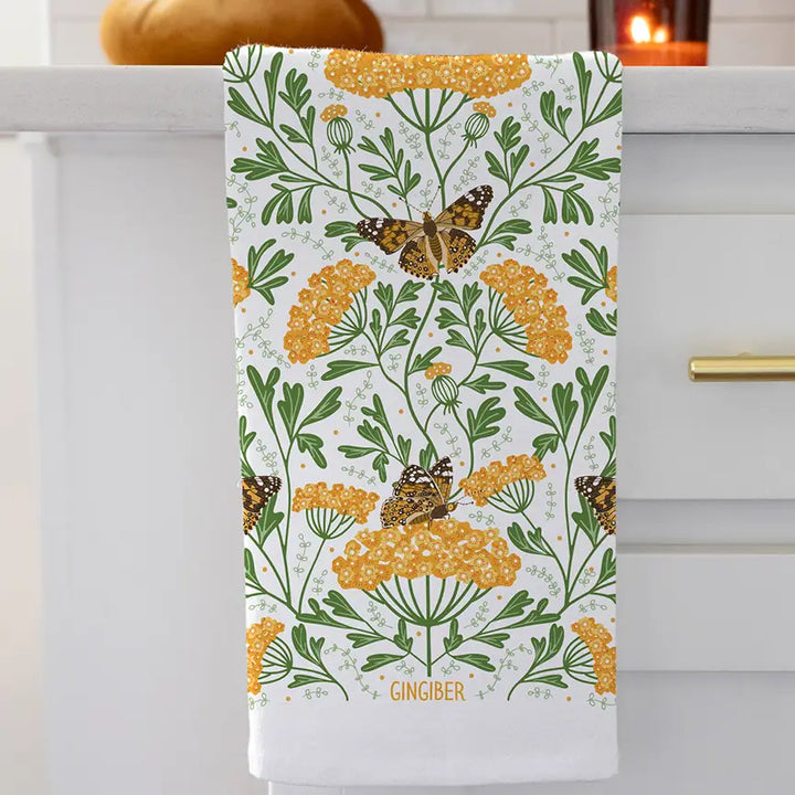 White cotton tea towel printed with a botanical pattern of golden yarrow flowers and painted lady butterflies, accented with green leaves hanging from a sink.